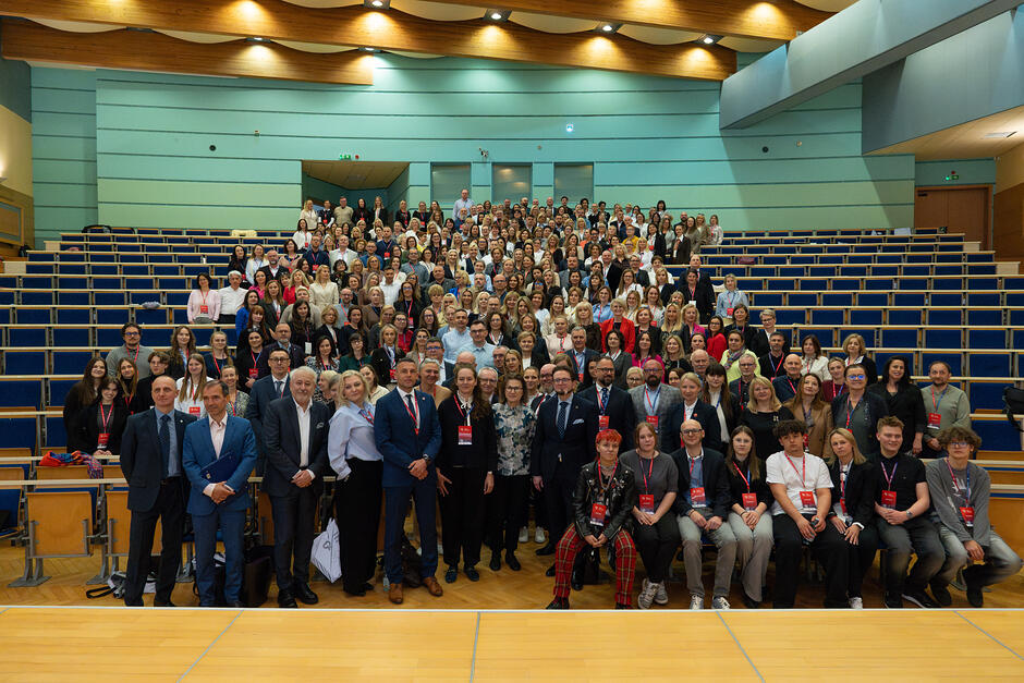 Zdjęcie przedstawia liczną grupę osób pozujących do wspólnej fotografii w dużej, nowoczesnej auli wykładowej z charakterystycznymi niebieskimi ścianami i drewnianym wykończeniem. Uczestnicy, ubrani głównie w stroje biznesowe i wyposażeni w identyfikatory, wypełniają zarówno przestrzeń przed sceną, jak i rzędy siedzeń wznoszące się ku tyłowi sali.
