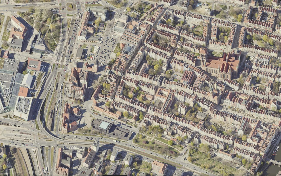 Zdjęcie przedstawia widok z lotu ptaka na gęsto zabudowane centrum miasta z charakterystycznymi kamienicami o czerwonych dachach oraz dużym, ceglanym kościołem. W lewej części kadru widoczna jest rozbudowana infrastruktura drogowa z wielopasmowymi jezdniami, torowiskami i nowoczesnymi budynkami użytkowymi.