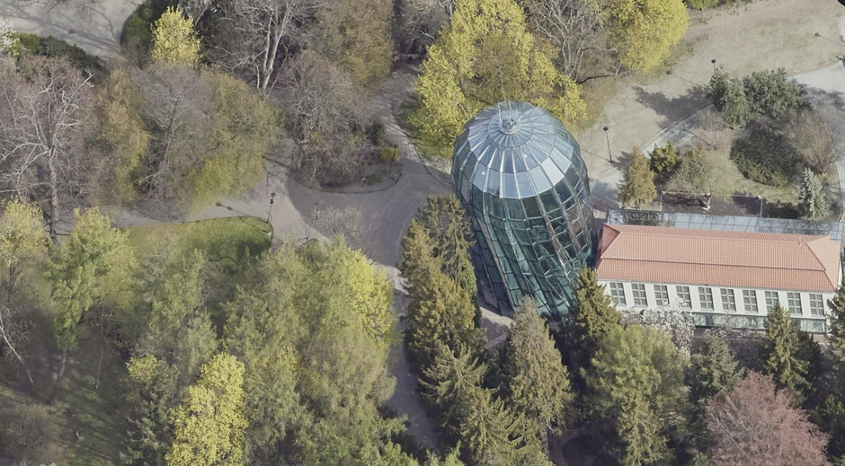 Zdjęcie przedstawia widok z lotu ptaka na nowoczesną, przeszkloną konstrukcję palmiarni o obłym kształcie, która sąsiaduje z podłużnym budynkiem o czerwonym dachu. Całość znajduje się w otoczeniu gęstego parku z licznymi drzewami o wiosennych barwach oraz wijącymi się alejkami spacerowymi.