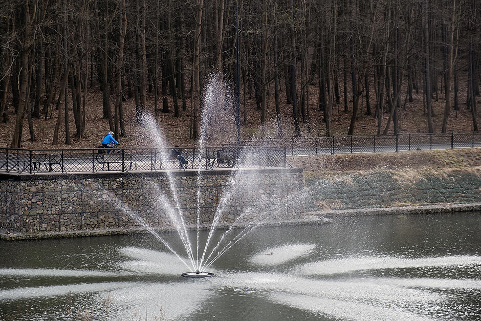 Fontanna na zbiorniku retencyjnym. Na jego brzegu siedzi osoba na ławce, obok jedzie osoba na rowerze.