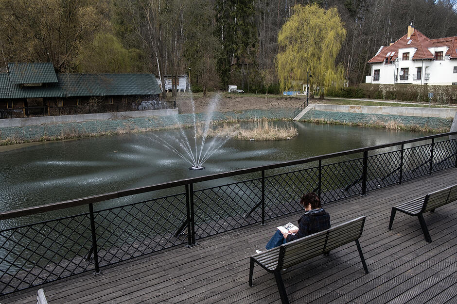 Nad zbiornikiem retencyjnym siedzi osoba na ławce i czyta książkę. Na stawie tryska woda z fontanny. W tle las.