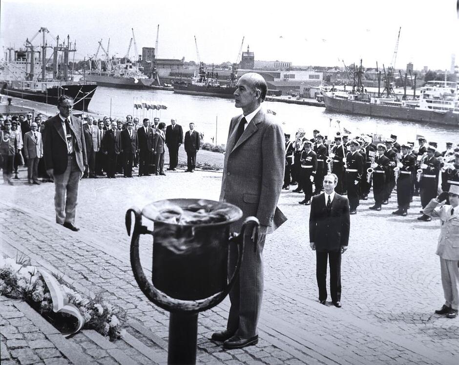 Szczupły mężczyzna w garniturze stoi samotnie oddając hołd - w oddaleniu kilku metrów wokół widać inne osoby 