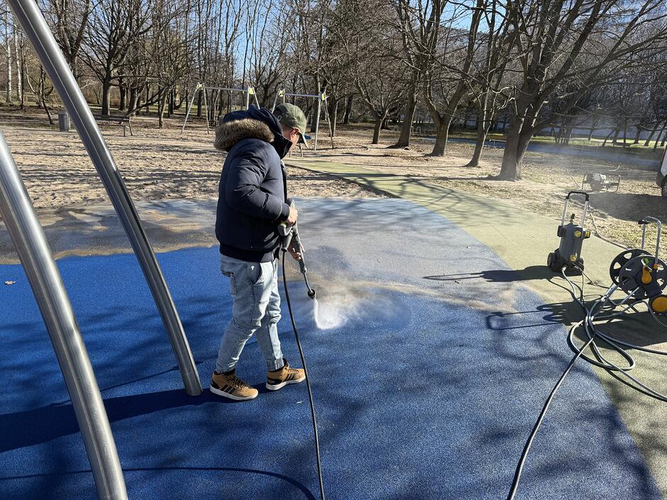 na zdjęciu mężczyzna w ręku z wężem z wodą, sprząta nawierzchnie placu zabaw