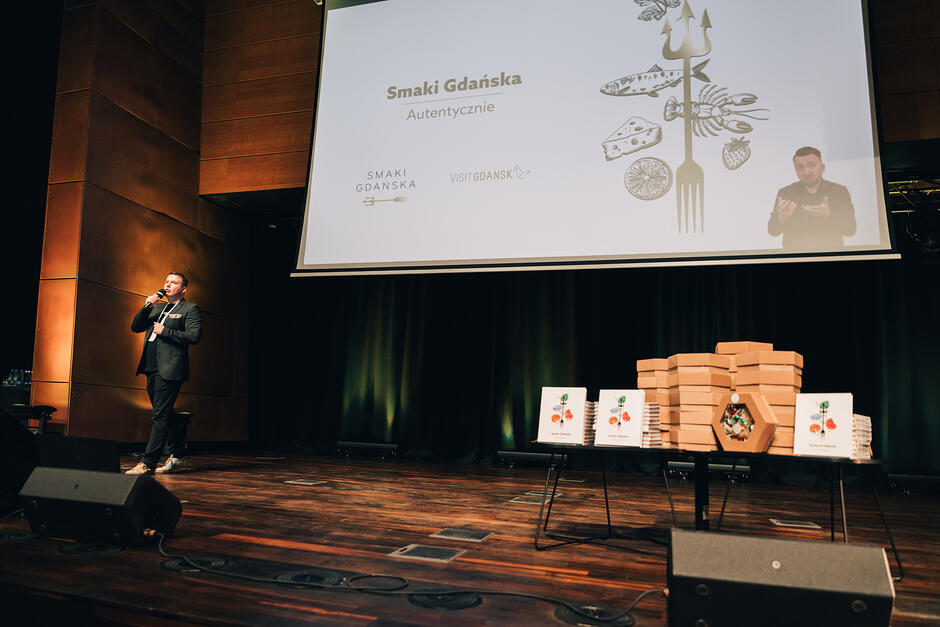 Mężczyzna przemawia na scenie w sali konferencyjnej, nad nim ekran z prezentacją.