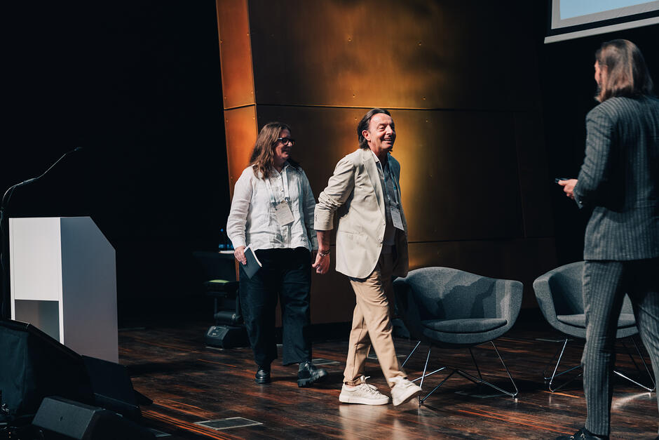 Mężczyzna idzie po scenie w sali konferencyjnej i śmieje się, inny mężczyzna go wita na scenie.