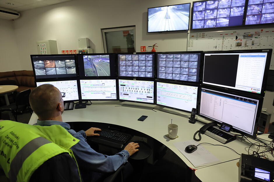 Mężczyzna w seledynowej kamizelce siedzi przed pulpitem z wieloma ekranami.