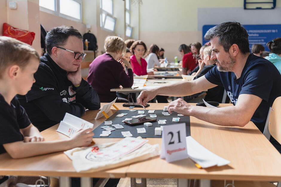 Zdjęcie przedstawia grupę osób w różnym wieku, które w skupieniu grają w grę planszową przy długich stołach. Uczestnicy, wśród których są dorośli i dzieci, rywalizują w jasnej sali, co sugeruje odbywający się właśnie turniej lub spotkanie miłośników gier.