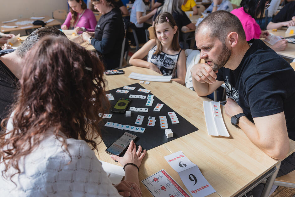 Zdjęcie ukazuje grupę ludzi, w tym mężczyznę i dziewczynkę, grających wspólnie w Rummikuba przy jasnym, drewnianym stole. Atmosfera wydaje się być pełna skupienia, a widoczne na stole tabliczki z numerami sugerują, że są to zorganizowane rozgrywki turniejowe.