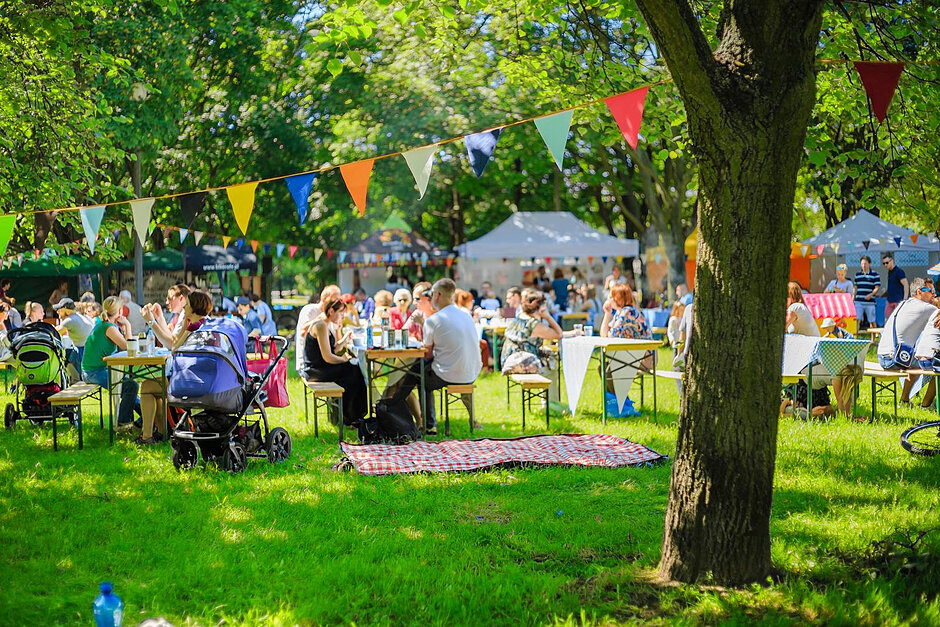 Ludzie siedzą przy stolikach na zielonej trawie. W tle piknik.