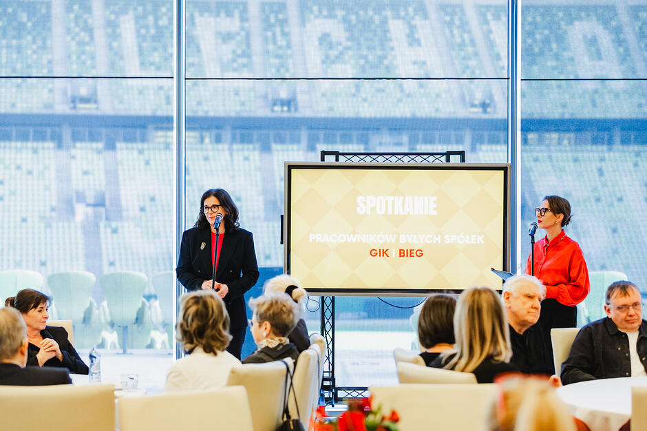 W sali konferencyjnej dwie kobiety stoją przy mikrofonach, przy okrągłych stołach siedzą ludzie. Za oknami widać trybuny stadionu.