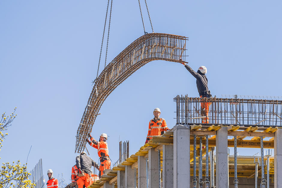 plac budowy, na budowanym budynku stoi kilku pracowników w roboczych kombinezonach, łapią duży metalowy element