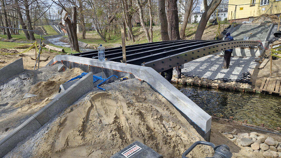 na zdjęciu metalowa konstrukcja nowego mostku, w tle park z drzewami