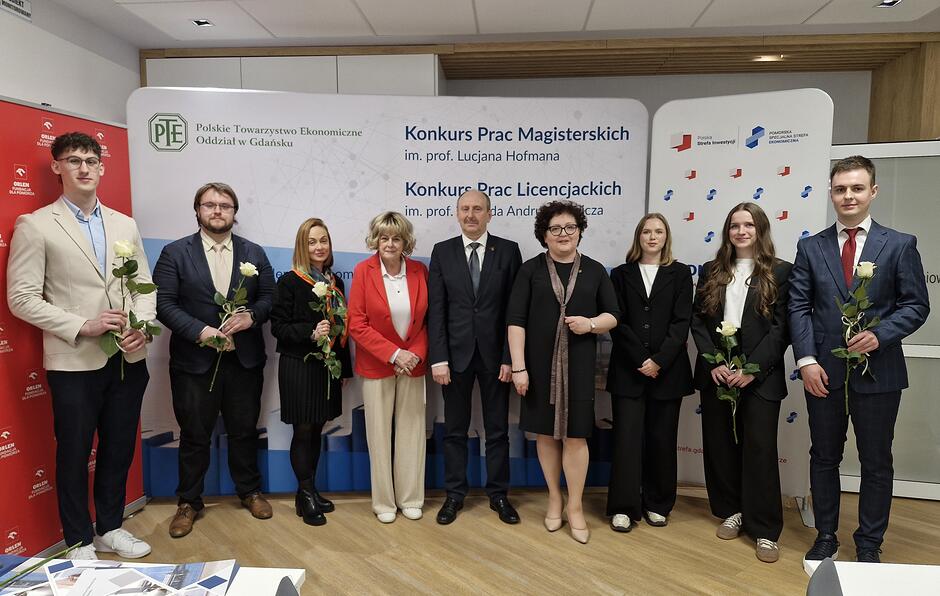 dziewięć osób pozuje do wspólnego zdjęcia na tle tzw. ścianek  z nazwami instytucji. Piec osób trzyma w ręku białą różę. 