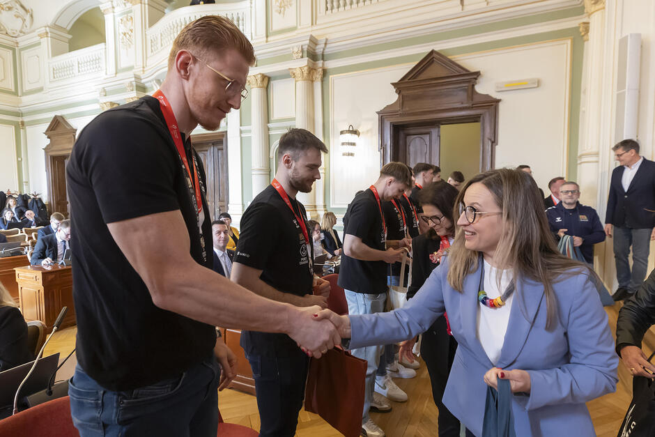 Kobieta w niebieskiej marynarce po prawej podaje dłoń wysokiemu mężczyźnie po lewej