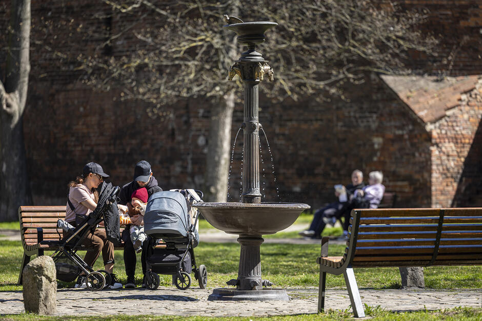 Ludzie siedzą na ławkach przy fontannie na miejskim skwerku.