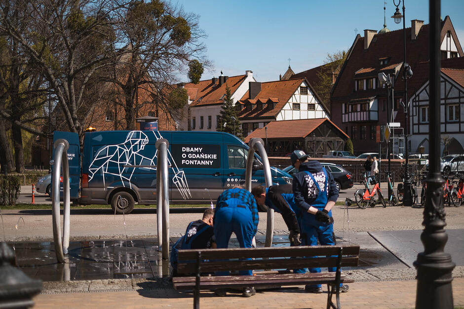 Dwaj mężczyźni pracują przy wyłączonej fontannie, w tle samochód bus z napisem dbamy o gdańskie fontanny .