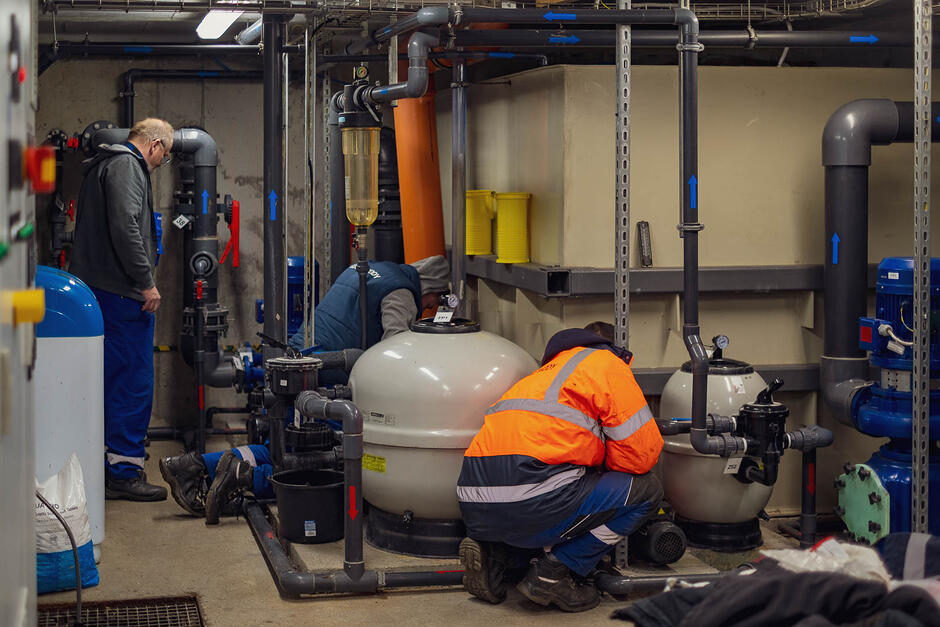 Pomieszczenie pełne rur, zaworów i zbiorników na wodę, w którym pracuje kilka osób w kombinezonach. 