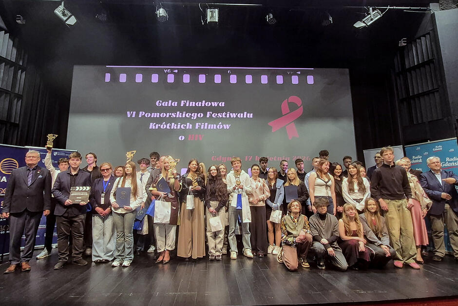 Duża grupa ludzi stoi na scenie w sali kinowej i pozuje do zdjęcia. Część z nich trzyma w rękach nagrody.