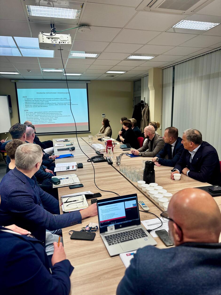 na zdjęciu kilkanaście dorosłych osób siedzi przy stole, patrzą na telebim