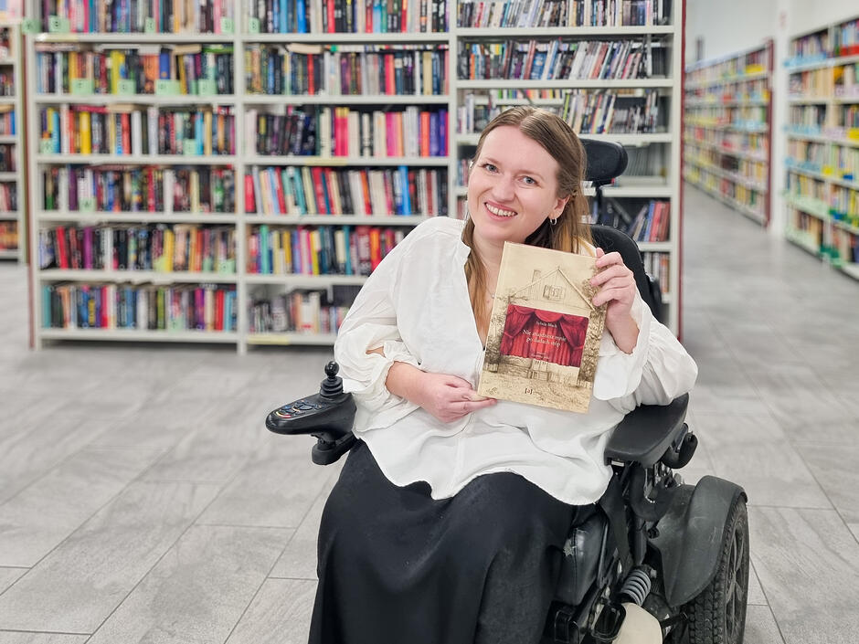 Osoba poruszająca się na wózku elektrycznym siedzi w jasnej, przestronnej bibliotece. W tle znajdują się wysokie regały wypełnione książkami. Osoba trzyma książkę z ilustracją teatralnej kurtyny na okładce.
