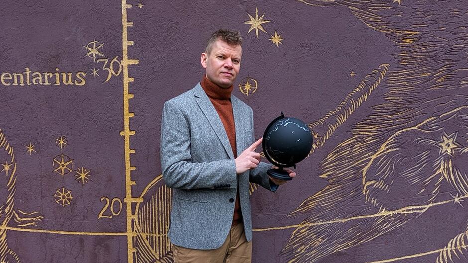 Osoba stoi na tle ściany ozdobionej ilustracją z motywami astronomicznymi, takimi jak gwiazdy, linie i oznaczenia przypominające mapę nieba. W dłoniach trzyma czarny globus, wskazując na niego palcem. Osoba ubrana jest w marynarkę i golf, a cała scena znajduje się na zewnątrz, przy dekoracyjnej elewacji budynku.