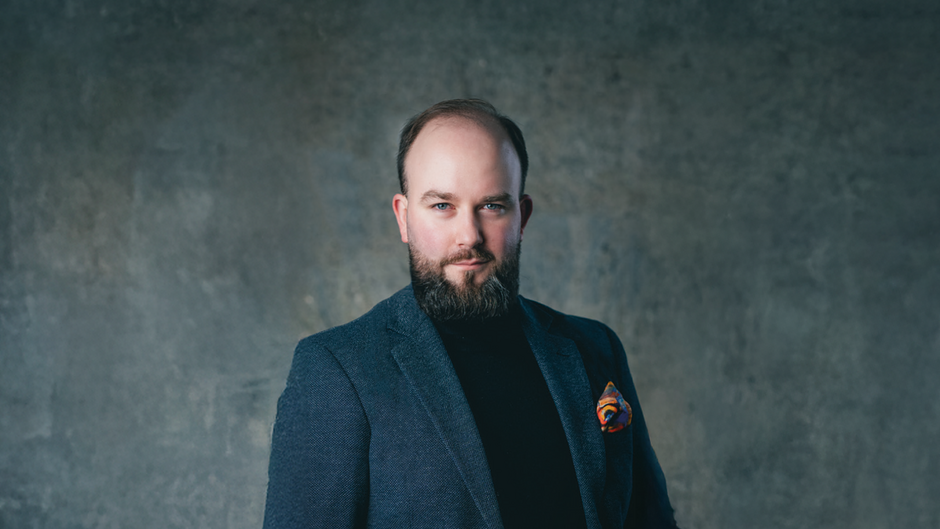 Portret studyjny osoby ujętej od pasa w górę, stojącej na tle jednolitej, szaro-niebieskiej ściany. Osoba ma krótkie włosy oraz zarost i ubrana jest w ciemną marynarkę założoną na gładki golf. W kieszonce marynarki widoczna jest kolorowa poszetka. Oświetlenie jest miękkie i skoncentrowane na sylwetce, tło pozostaje lekko rozmyte.