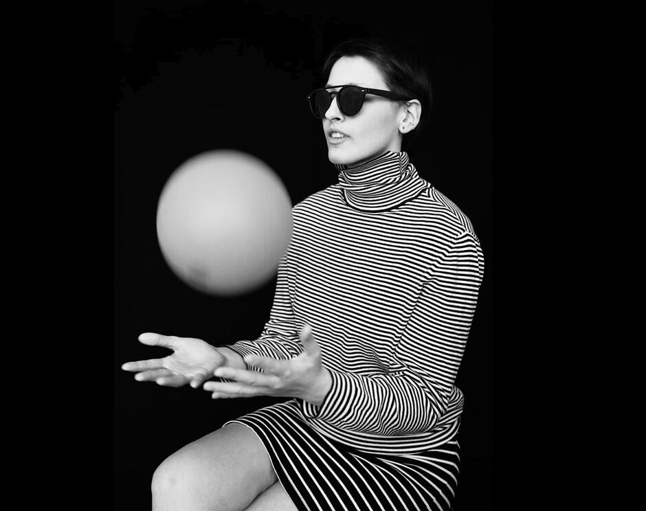 Czarno-białe zdjęcie osoby siedzącej na tle jednolitego, ciemnego tła. Osoba ma krótkie włosy i nosi ciemne okulary oraz golf w poziome paski. Dłonie są uniesione przed sylwetką, a pomiędzy nimi unosi się jasna, okrągła kula. Osoba siedzi bokiem do kadru, a cała scena ma minimalistyczny, studyjny charakter z wyraźnym kontrastem światła i cienia.