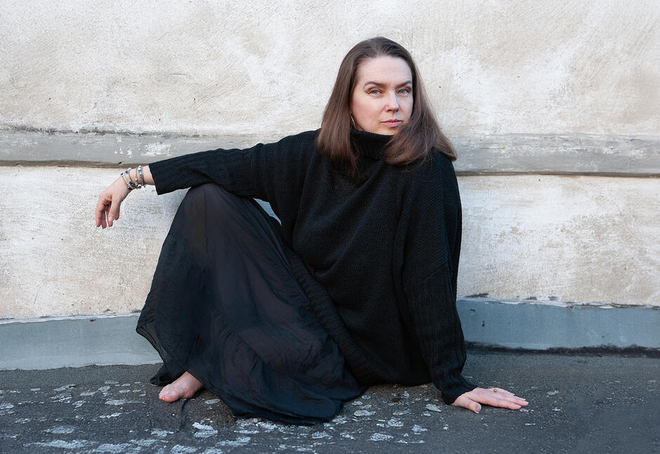 Osoba siedzi na ziemi przy jasnej, lekko zniszczonej ścianie budynku. Jedna noga jest podkurczona, druga wyciągnięta, a jedna dłoń oparta o podłoże. Osoba ubrana jest w czarną, luźną bluzę z długim rękawem oraz długą, ciemną spódnicę. Widoczne są bransoletki na nadgarstku. Zdjęcie wykonane jest w naturalnym świetle, z prostym, minimalistycznym tłem.