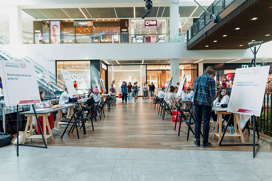 W galerii handlowej stoją rozstawione stanowiska gdzie można zrobić badania. Za stołami siedzą osoby w białych fartuchach. Pomiędzy chodzą ludzie.