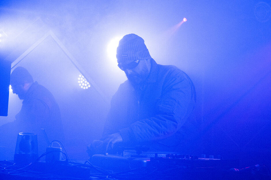 Osoba stojąca przy stole DJ‑skim obsługuje mikser i kontrolery podczas występu na scenie, oświetlonej intensywnym niebieskim światłem i wypełnionej dymem; w tle widoczna druga postać oraz reflektory sceniczne.