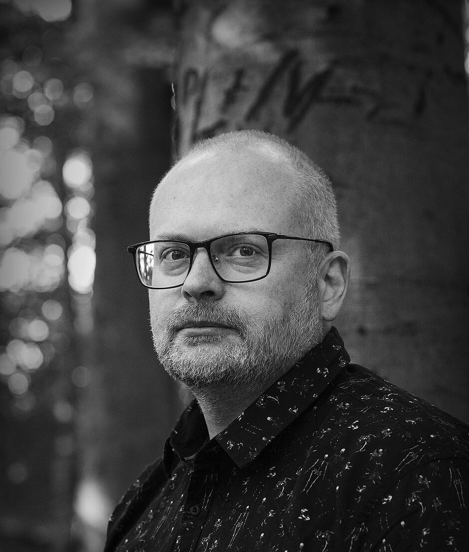 Czarno-biały portret osoby stojącej na zewnątrz; widoczna głowa i ramiona na tle pnia drzewa oraz rozmytego leśnego tła. Osoba ma krótkie włosy i okulary, ubrana jest w ciemną koszulę z jasnym, nieregularnym wzorem. 