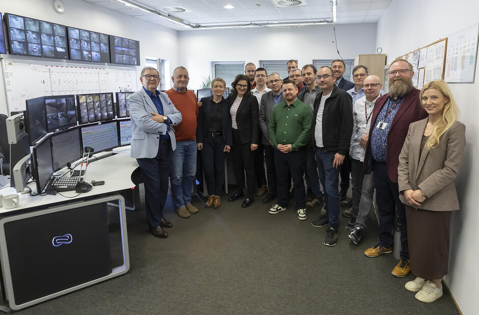 Grupa kilkunastu osób pozuje do wspólnej fotografii w pokoju z wieloma monitorami na długim stole i na ścianach.