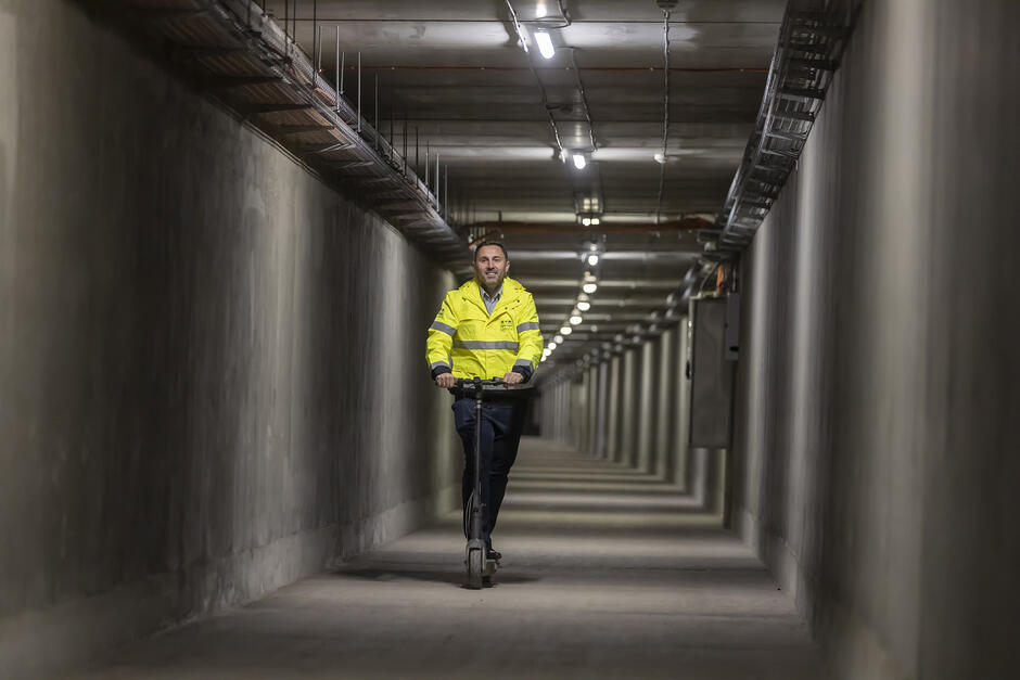 Mężczyzna w seledynowej kurtce jedzie na hulajnodze przez tunel.