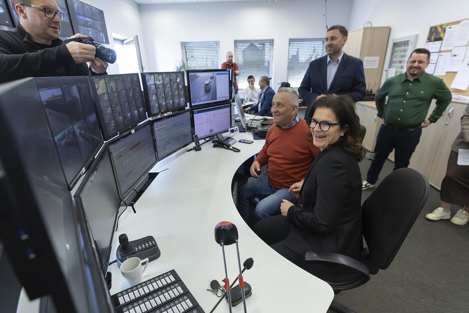 W pokoju z długim stołem i wieloma monitorami jest kilka osób. Kobieta i mężczyzna siedzą przy stole, patrzą w monitory i śmieją się. Wokół stoi kilka innych osób.