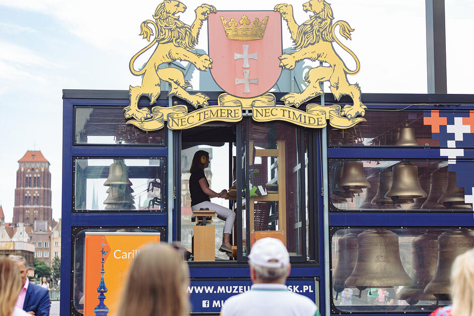 Zdjęcie przedstawia mobilny carillon z herbem Gdańska, na którym artystka gra na zestawie dzwonów w otoczeniu publiczności. W tle widać charakterystyczną sylwetkę Bazyliki Mariackiej, co podkreśla miejski i historyczny klimat tego muzycznego wydarzenia.
