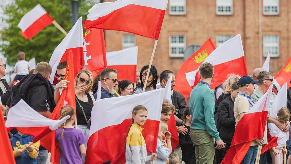 Zdjęcie przedstawia tłum ludzi uczestniczących w patriotycznym zgromadzeniu, nad którym powiewa wiele biało-czerwonych flag Polski. Wśród uczestników w różnym wieku widać zarówno dorosłych, jak i dzieci, a w tle znajdują się miejskie budynki z cegły.