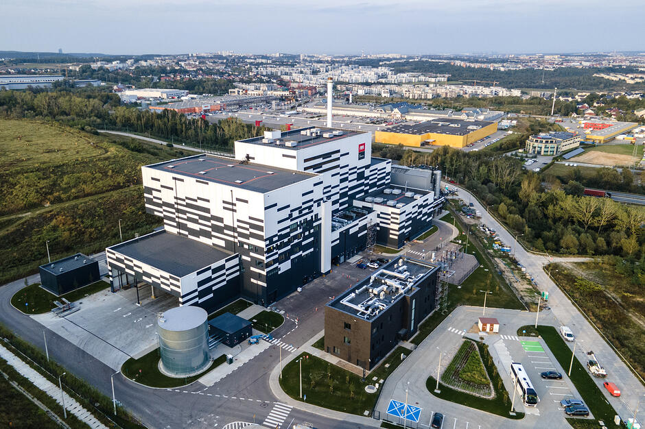 Zdjęcie przedstawia nowoczesny, wielkogabarytowy budynek przemysłowy o biało-czarnej elewacji, który jest siedzibą Portu Czystej Energii w Gdańsku. Obiekt otoczony jest infrastrukturą drogową i parkingami, a w oddali rozciąga się panorama miasta z licznymi zabudowaniami i terenami zielonymi.