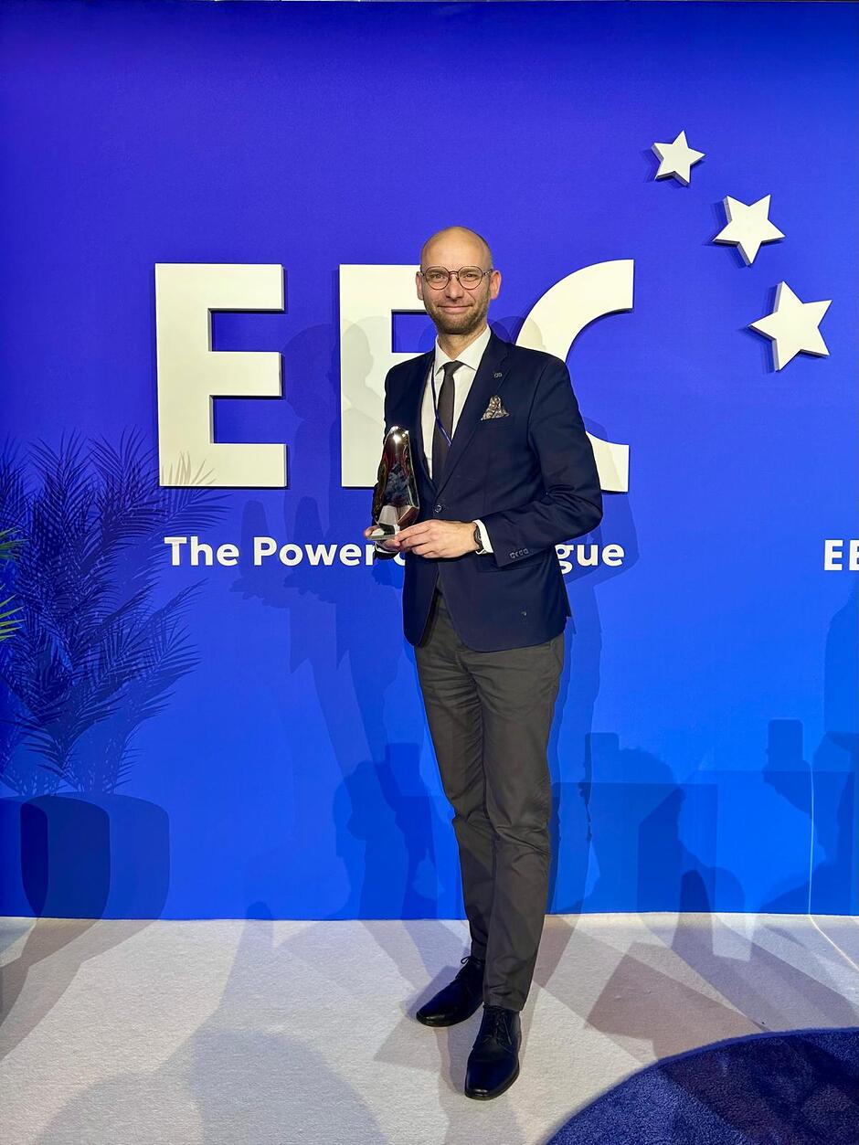 Zdjęcie przedstawia uśmiechniętego mężczyznę w garniturze, który trzyma srebrną statuetkę na tle niebieskiej ścianki z logo Europejskiego Kongresu Gospodarczego (EEC). Całość dokumentuje uroczysty moment wręczenia nagrody podczas gali odbywającej się w ramach tego prestiżowego wydarzenia biznesowego.