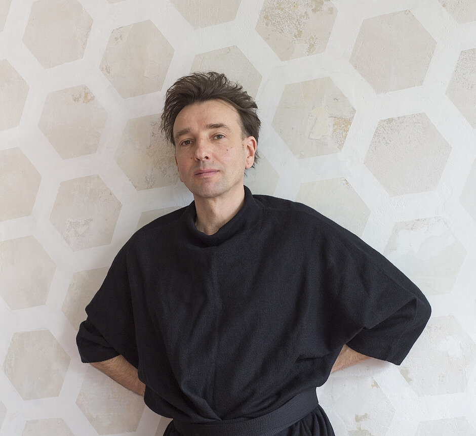 Osoba dorosła stoi na jasnym tle z geometrycznym wzorem przypominającym sześciokąty. Ubrana jest w luźny, czarny strój z krótkimi rękawami, przewiązany paskiem w talii. Ramiona są cofnięte, dłonie schowane za plecami. Włosy są krótkie, ciemne i nieco uniesione. Postać ustawiona jest frontalnie, lekko pochylona w jedną stronę, na tle jasnej, fakturowanej ściany.