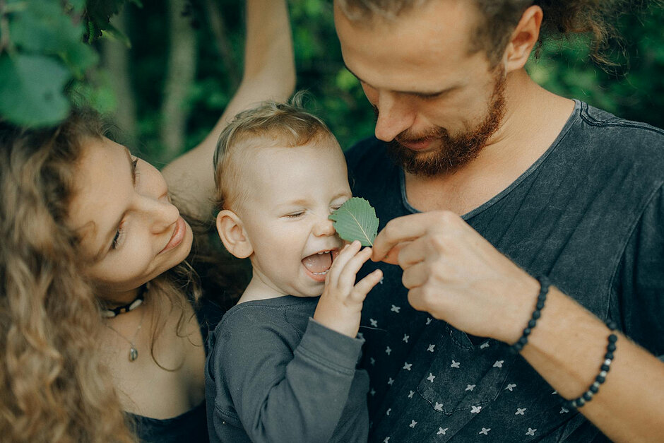 Młodzi kobieta i mężczyzna z dzieckiem pośród zieleni. Mężczyzna dotyka nosa dziecka liściem, te się śmieje.