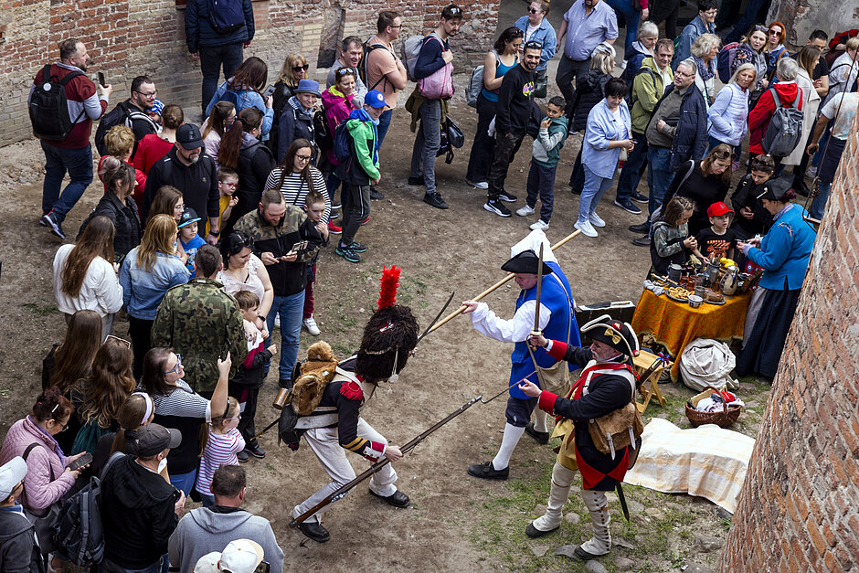 Trzej żołnierze w mundurach z dawnej epoki walczą na bagnety i włócznie na dziedzińcu starej ceglanej budowli. Otacza ich tłum widzów.