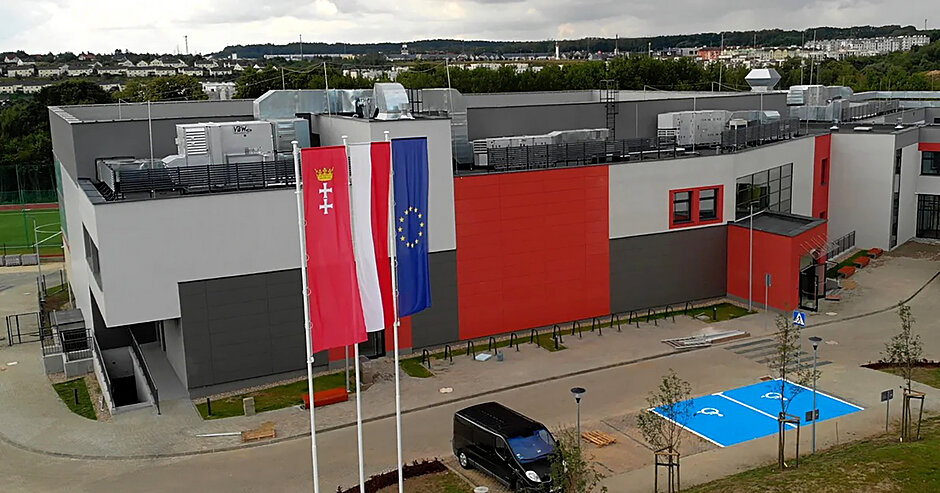 Zdjęcie z lotu ptaka, widok na budynek szkoły, przed którym na maszcie powiewają flagi Polski, Unii Europejskiej i Gdańska.