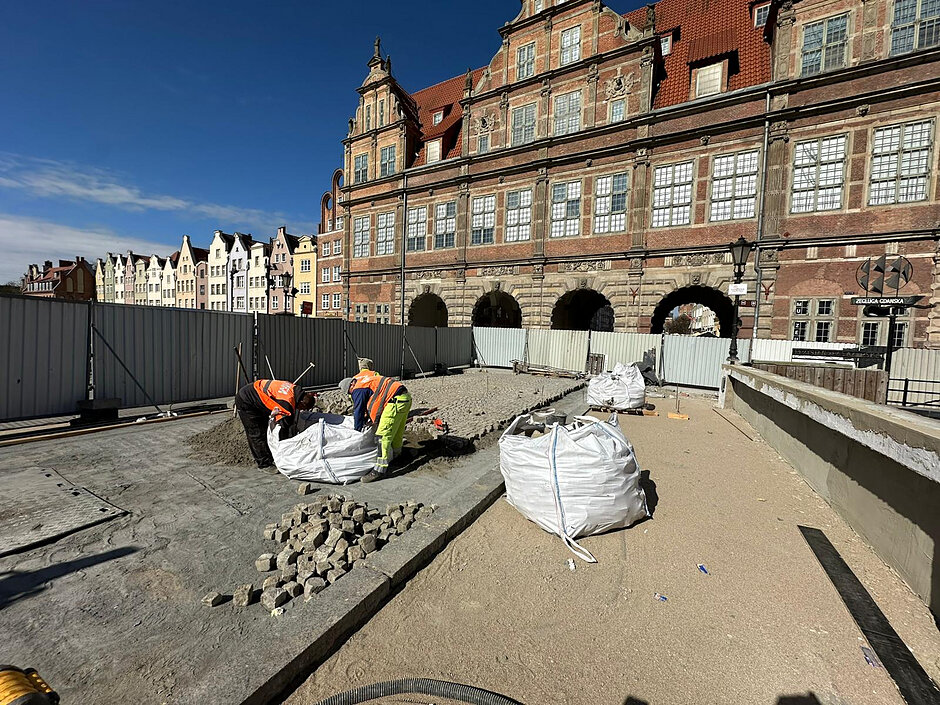 na zdjęciu dwóch ludzi układających na moście kostkę brukową, w tle zabytkowy ozdobny budynek