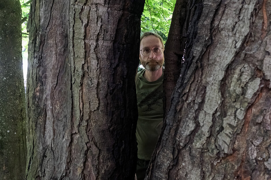 Brodaty mężczyzna w okularach spogląda w stronę aparatu przez wąską szczelinę między dwoma grubymi pniami drzew o wyraźnej teksturze kory. W tle widać soczystą zieleń liści, co sugeruje, że zdjęcie zostało wykonane w lesie lub parku.
