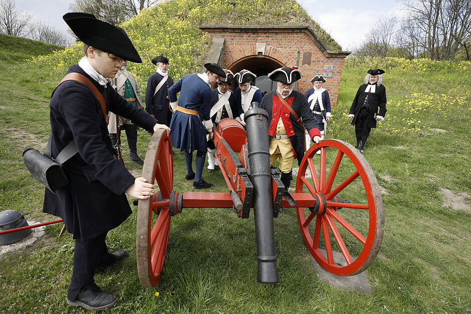 Zdjęcie przedstawia grupę rekonstrukcyjną w historycznych strojach wojskowych, która obsługuje czerwoną armatę na tle ceglanej budowli obronnej. Mężczyźni w trójgraniastych kapeluszach i ciemnych mundurach przygotowują działo do wystrzału na trawiastym terenie otoczonym żółtymi kwiatami.