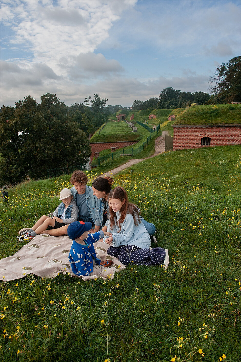 Zdjęcie przedstawia uśmiechniętą rodzinę z trójką dzieci, która spędza czas na pikniku na zielonym zboczu usianym żółtymi kwiatami. W tle widać zabytkowe, ceglane zabudowania obronne, co sugeruje, że wypoczywają oni na malowniczym terenie dawnych fortyfikacji.
