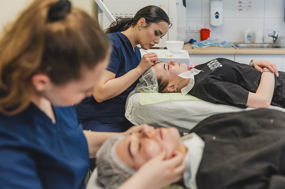 Zdjęcie zrobione podczas wykonywania zabiegów kosmetologicznych. Widać dwie kosmetyczki i dwie leżące osoby korzystające z ich usług. 