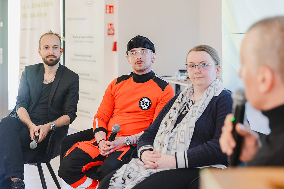 Trójka uczestników siedzi w rzędzie, przysłuchując się wypowiedzi osoby z mikrofonem, która znajduje się na pierwszym planie po prawej stronie. Wyróżniający się jaskrawopomarańczowy mundur jednego z mężczyzn wskazuje, że dyskusja może dotyczyć ratownictwa lub bezpieczeństwa publicznego.