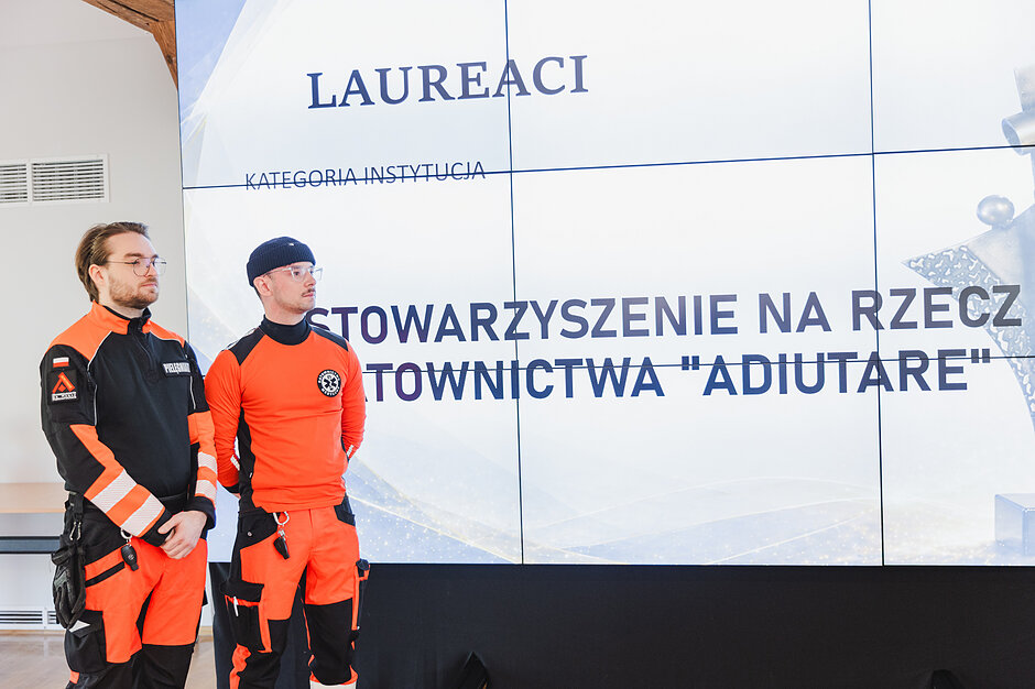 Dwaj mężczyźni w profesjonalnych strojach ratowników medycznych stoją przed dużym ekranem, na którym widnieje napis o laureatach w kategorii „Instytucja”. Treść na wyświetlaczu wskazuje, że wyróżnienie otrzymało Stowarzyszenie na rzecz Ratownictwa „Adiutare”.