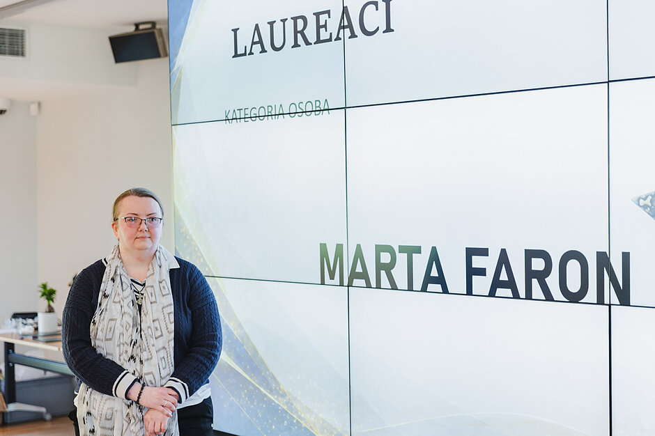 Kobieta w okularach i wzorzystym szalu stoi przed wielkoformatowym ekranem w jasnym pomieszczeniu. Wyświetlony na nim napis informuje o laureatce w kategorii „Osoba”, którą została Marta Faron.